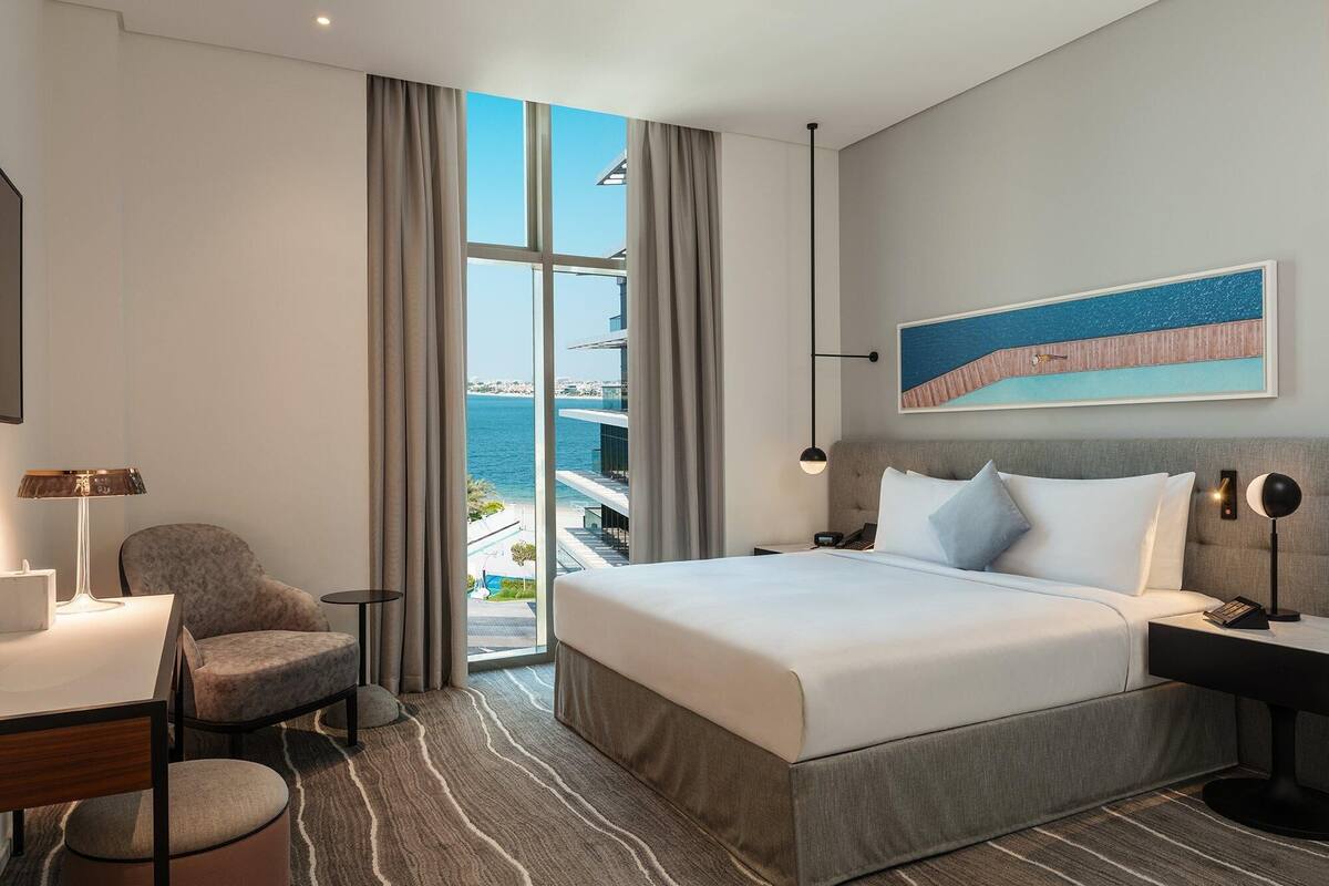 A hotel room with a large bed, a chair, a desk, and a view of the sea.