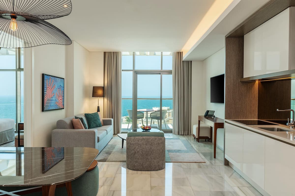 A modern living room with a sofa, coffee table, and ottoman, a dining area with a table and chairs, a kitchen with a sink and cabinets, and a large window with a view of the sea.