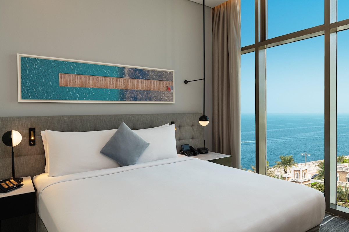 A hotel room with a large bed, a nightstand, a phone, a lamp, and a view of the ocean.