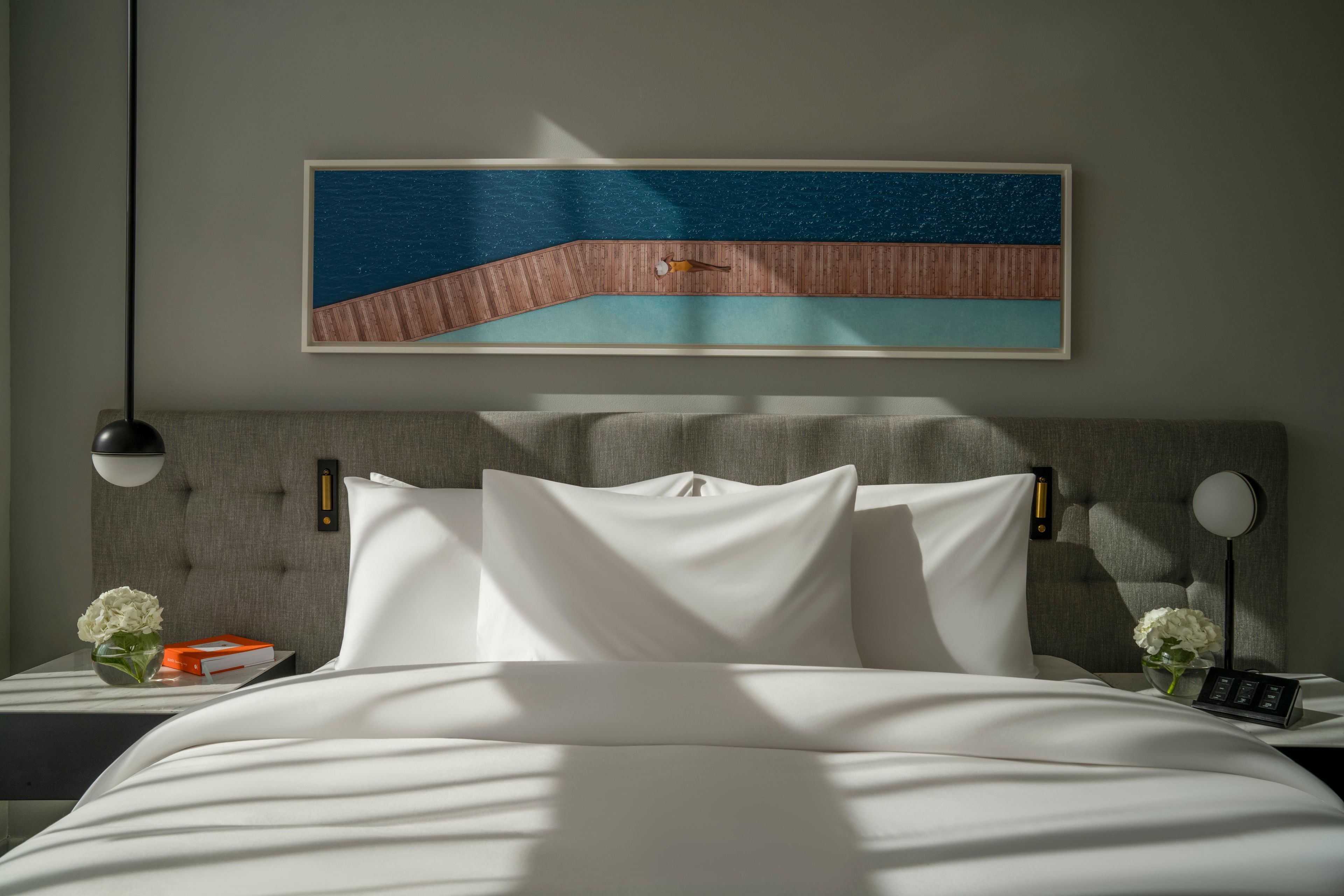 A neatly made bed with white linens, a headboard with a textured design, a framed artwork above the bed, and a bedside table with a vase of flowers and a book.