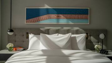 A neatly made bed with white linens, a headboard with a textured design, a framed artwork above the bed, and a bedside table with a vase of flowers and a book.