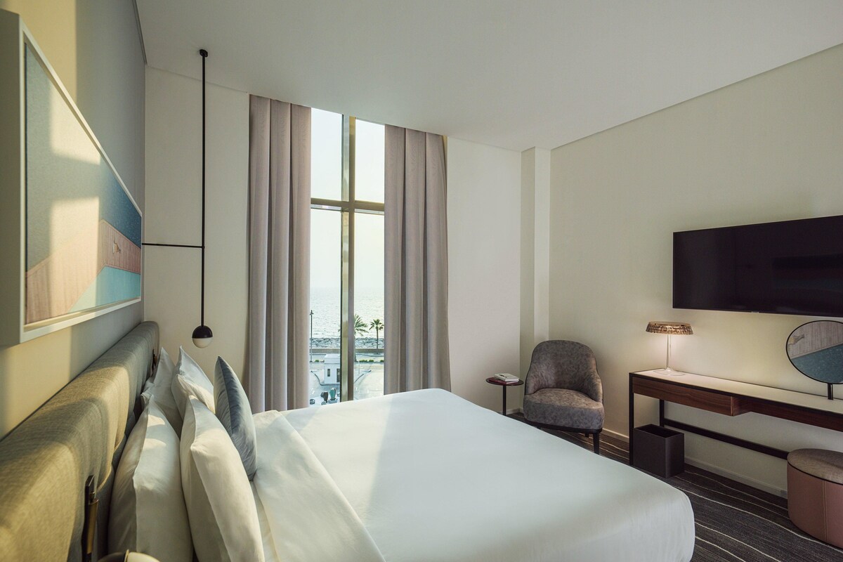 A hotel room with a large bed, a desk, a chair, a television, and a view of the sea.