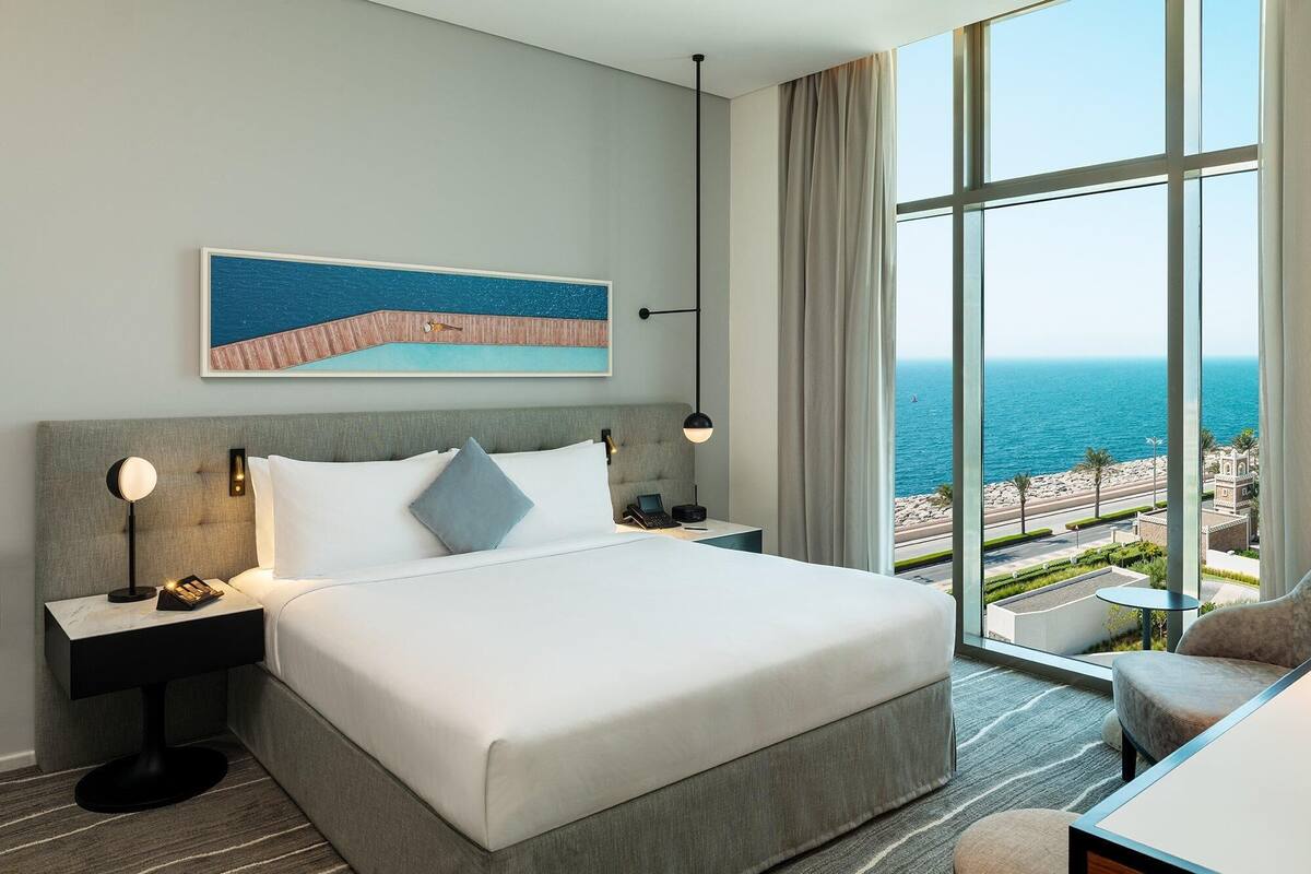 A hotel room with a large bed, a nightstand, a desk, and a view of the sea.