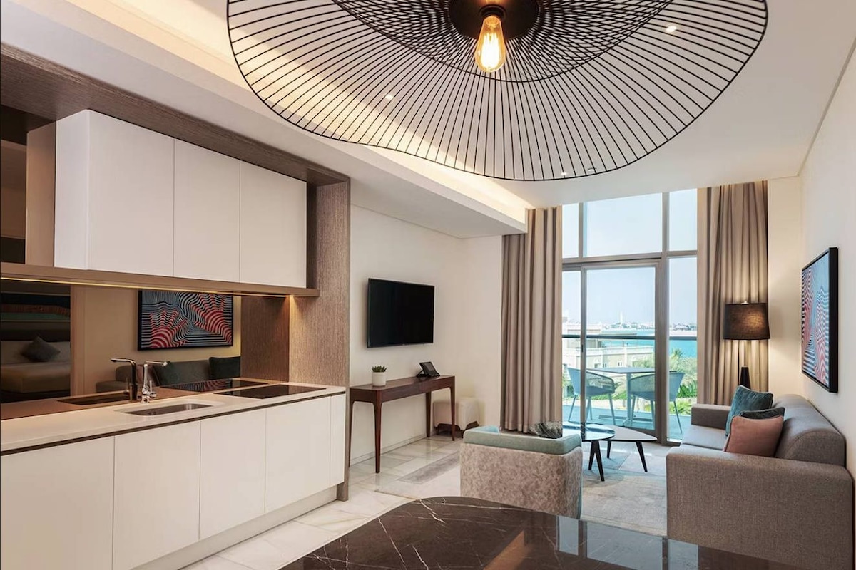 A modern kitchen with a marble countertop, a dining area with a glass table, a sofa, and a TV mounted on the wall.