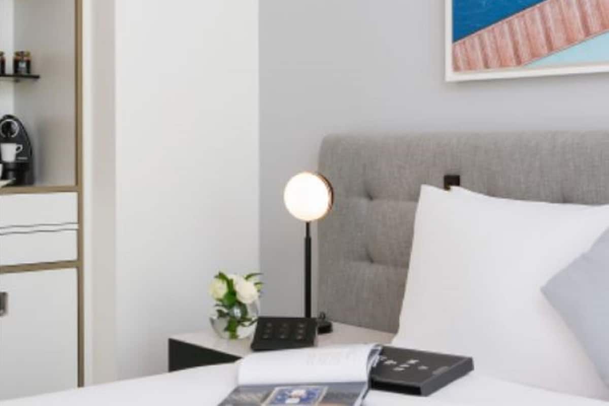 A neatly arranged hotel room with a bed, a bedside table with a lamp, a small vase with flowers, and a framed picture on the wall.
