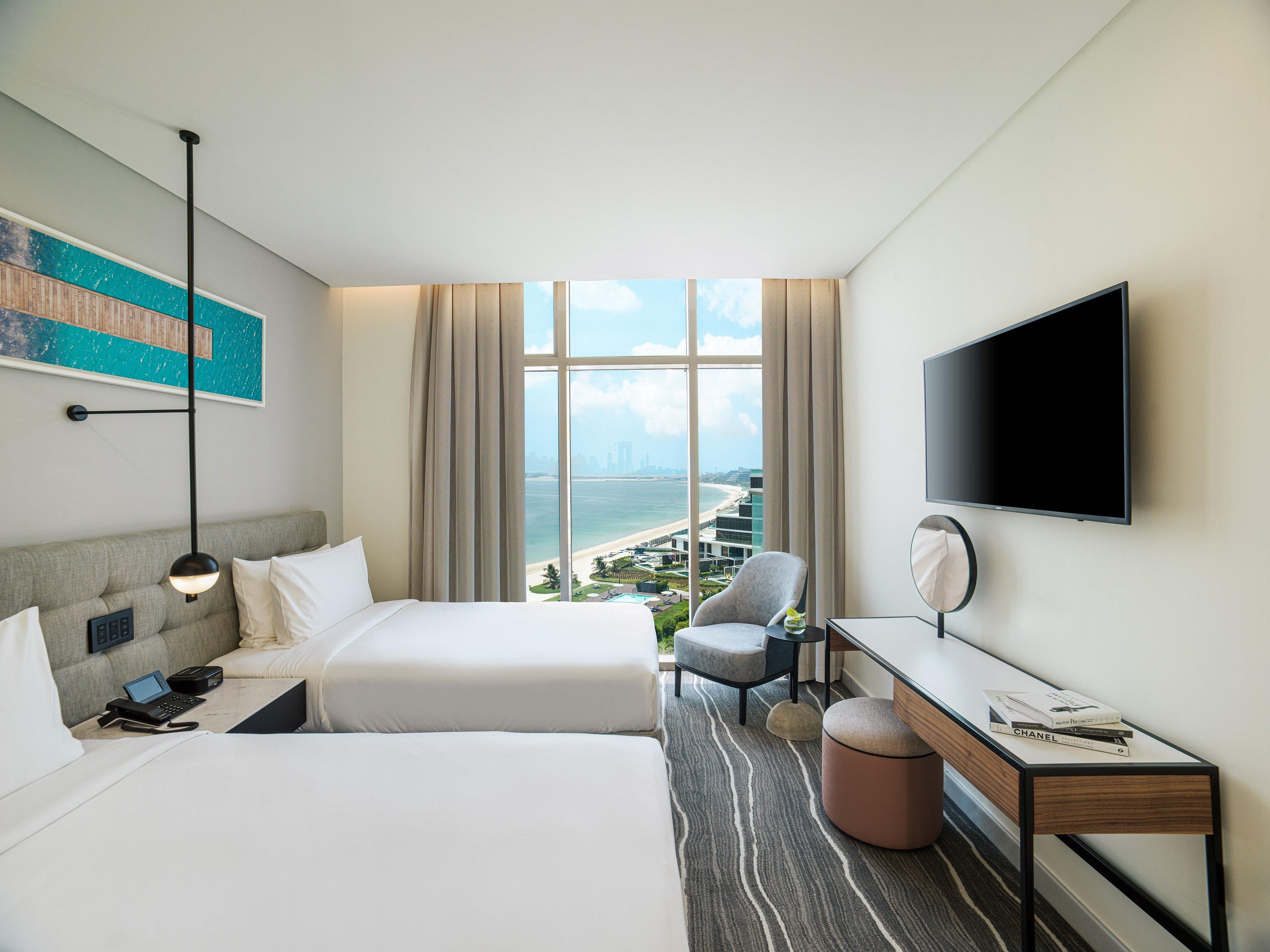 A hotel room with two beds, a desk, a chair, a TV, and a large window with a city view.