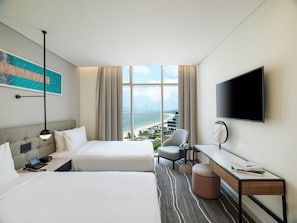A hotel room with two beds, a desk, a chair, a TV, and a large window with a city view. - Th8 Palm Dubai Beach Resort Vignette Collection (Dubai)