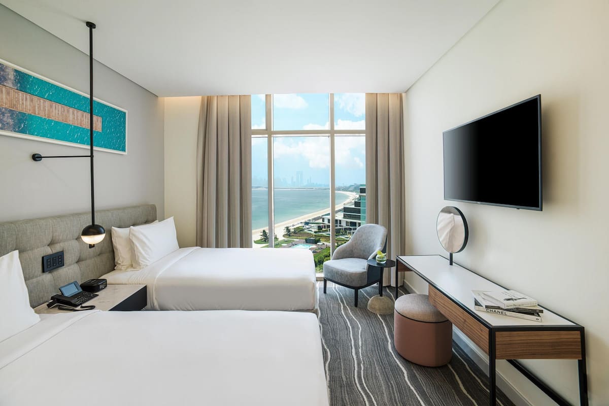 A hotel room with two beds, a desk, a chair, a TV, and a large window with a city view.