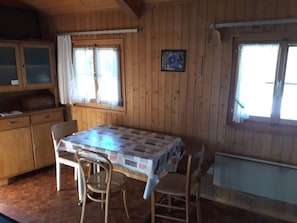 Ein Holzhütten-Interieur mit Esstisch, Stühlen, einem Schrank und zwei mit Vorhängen versehenen Fenstern.