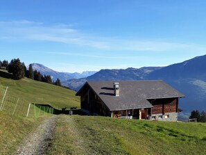 Una cabana de muntanya amb teulada inclinada, envoltada per una zona verda i un camí de terra.