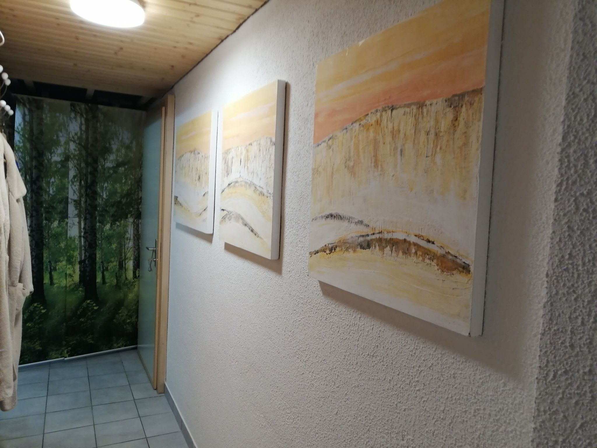 A hallway with three paintings on the wall, a door with a forest mural, and a tiled floor.