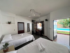 Quarto de hotel com duas camas, uma mesa de trabalho, uma cadeira, ventoinha no teto e vista para uma piscina.