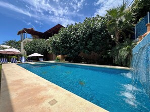 Una piscina con sillas de descanso y sombrillas, rodeada de exuberante follaje y un edificio con una cascada de azulejos.