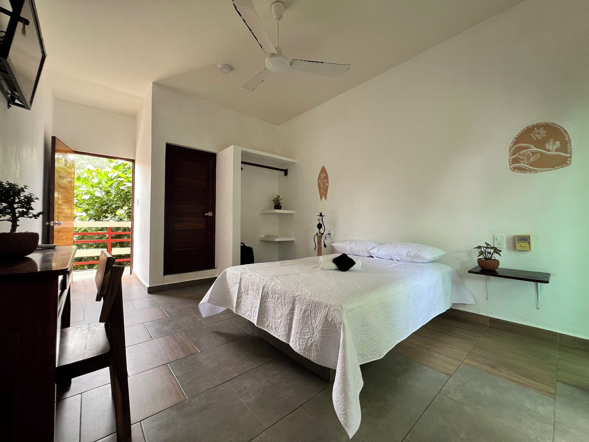 A bedroom with a bed, a desk, a chair, a ceiling fan, and a view of the outdoors.