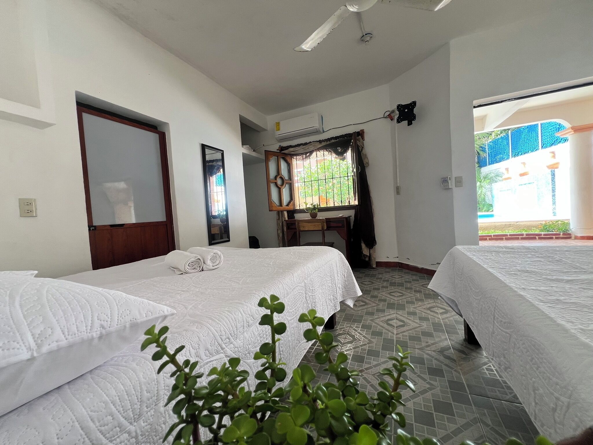 A hotel room with two beds, a desk, a mirror, and a window with a view of greenery.