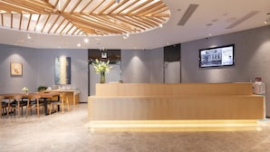 A modern reception area with a wooden reception desk, a dining area with chairs, a large abstract painting, and a wall-mounted television. - Hanting Hotel Shenzhen Nanshan Avenue Branch (Shenzhen)