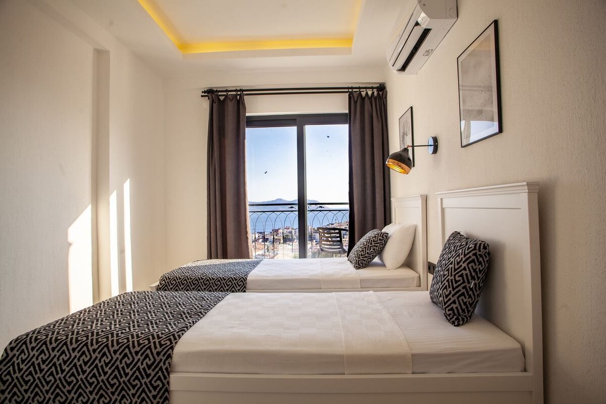 A hotel room with two beds, a large window with curtains, a view of the sea, and a ceiling light.