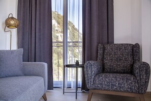 Habitación con sofá gris, sillón estampado, una mesita con planta y vistas amplias a través de ventanas con cortinas.