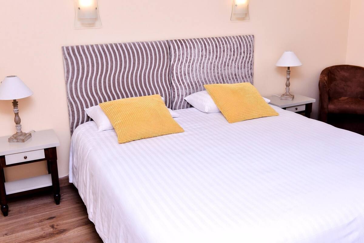 Una cama bien hecha con cabecera a rayas, dos lámparas de noche y una silla marrón.