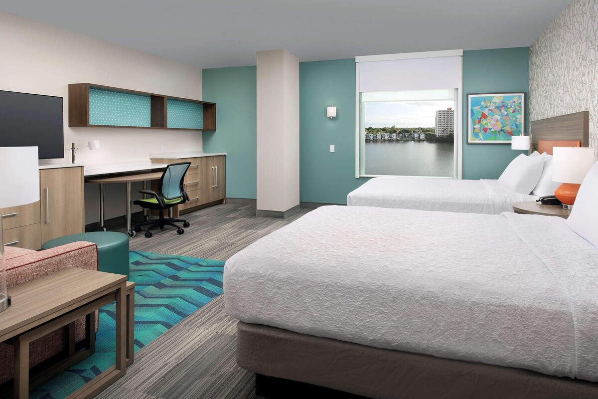 A hotel room with a bed, desk, chair, and a view of the city.
