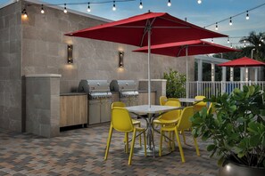 An outdoor patio with a brick floor, a barbecue grill, yellow chairs, and red umbrellas. - Home2suites by Hilton Miami Airport S. Blue Lagoon (Miami)