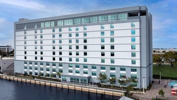 A modern hotel with a white facade, multiple floors, and a pool area with palm trees.