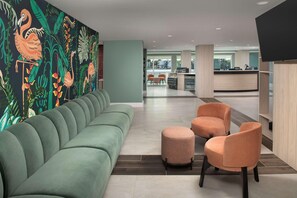 Un hall d’entrée moderne avec un canapé vert, des fauteuils orange, un comptoir d’accueil et une fresque représentant des plantes tropicales.