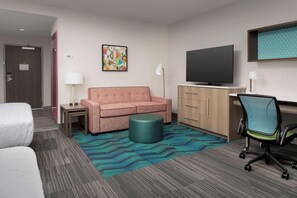 Une chambre d’hôtel comprenant un canapé, un pouf rond, un bureau avec une chaise, une télévision et un tapis à motifs.