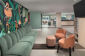 A modern lobby with a green sofa, orange chairs, a reception desk, and a mural with tropical plants. - Home2suites by Hilton Miami Airport S. Blue Lagoon (Miami)