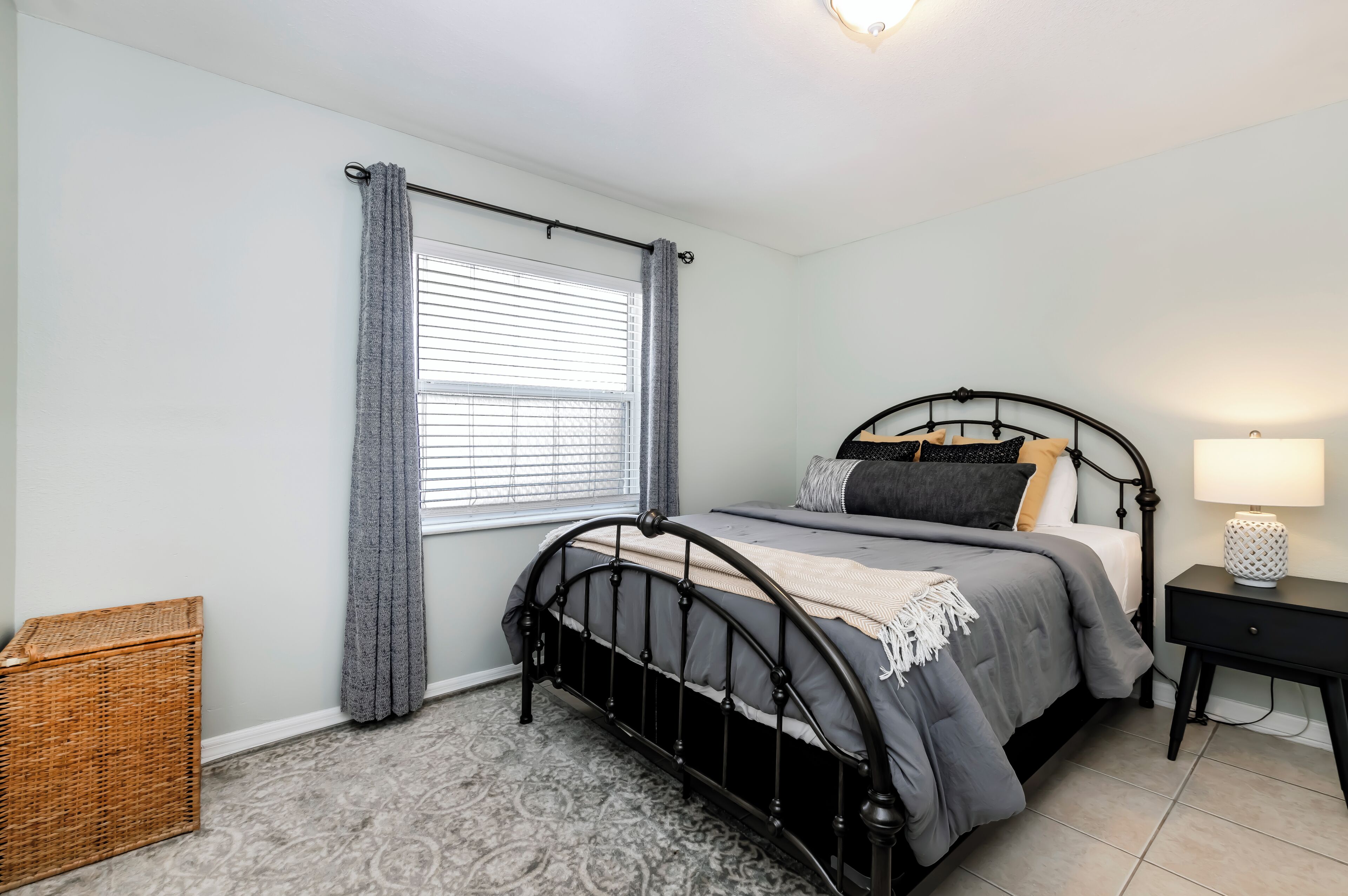 Quarto com cama, mesa de cabeceira, luminária, janela com cortinas e cesto de vime.