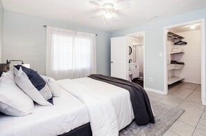 Ein Schlafzimmer mit einem Bett, einem Nachttisch, einem Deckenventilator und einem durchgegangenen Türspalte sichtbaren Badezimmer.