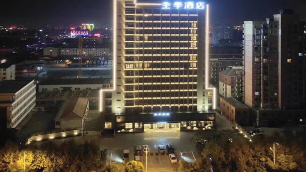 Um hotel com vários andares, um letreiro iluminado em destaque, área de estacionamento e edifícios urbanos ao redor.