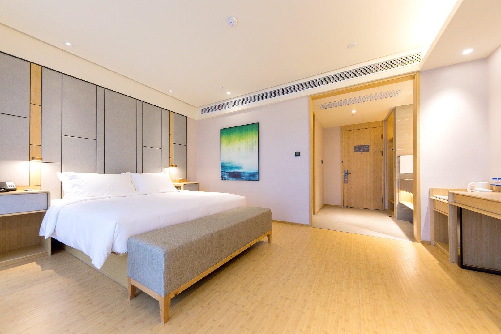 A hotel room with a bed, bedside table, bench, artwork, and a door leading to a bathroom.
