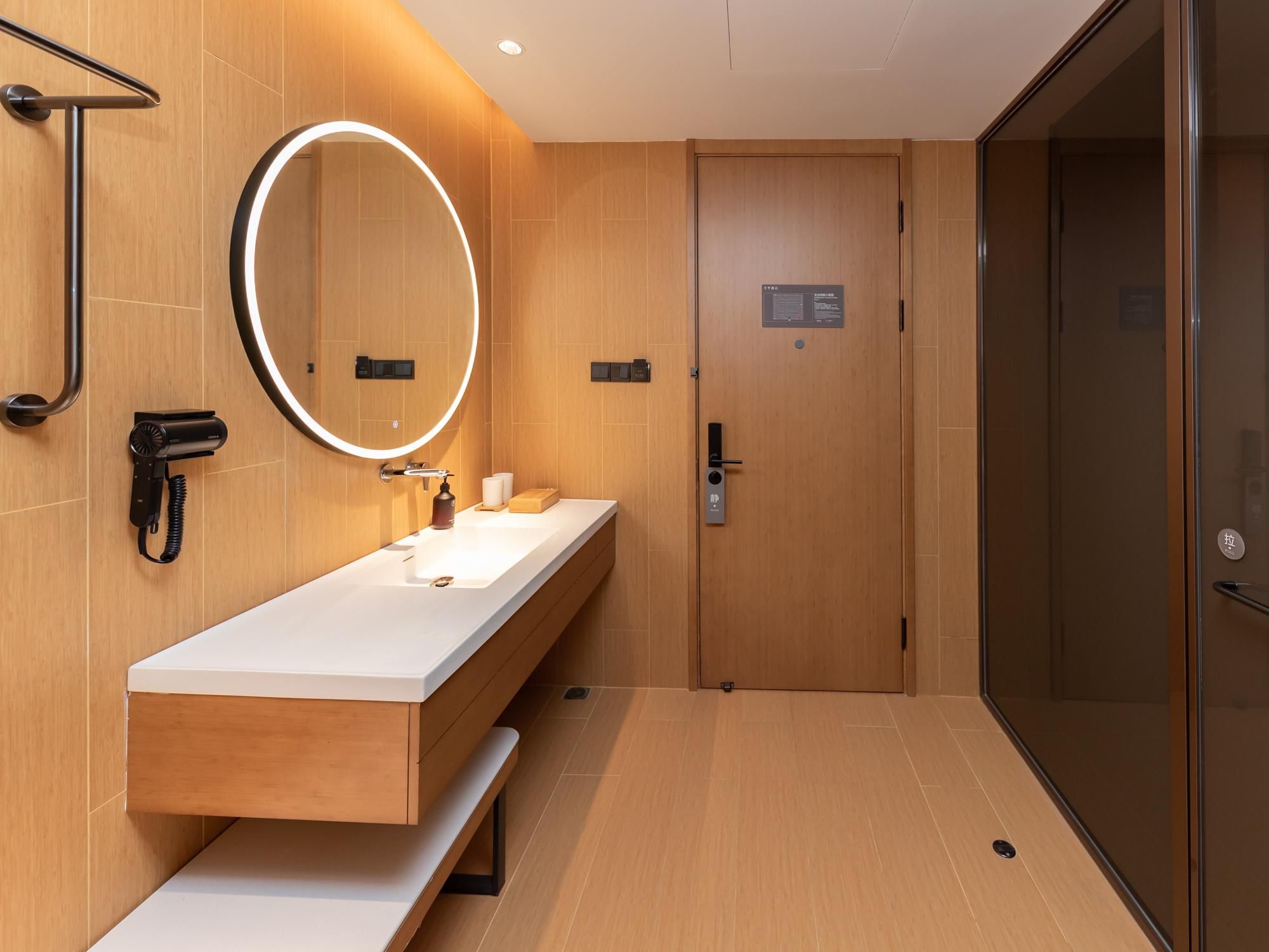 A modern bathroom with a wooden vanity, a mirror with a circular light, a towel dispenser, and a glass shower enclosure.