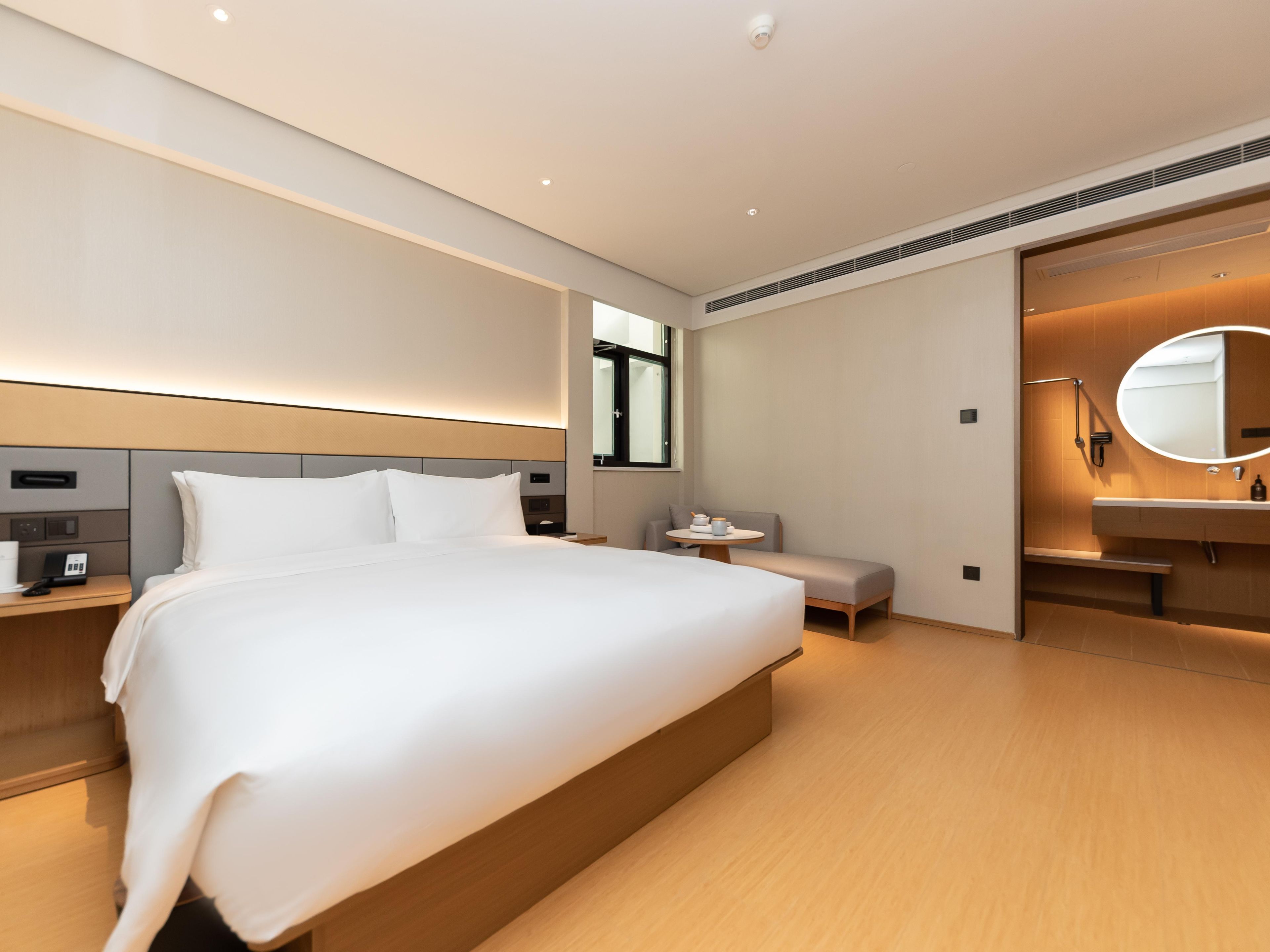 A modern hotel room with a large bed, bedside tables, a round mirror, and a small seating area.
