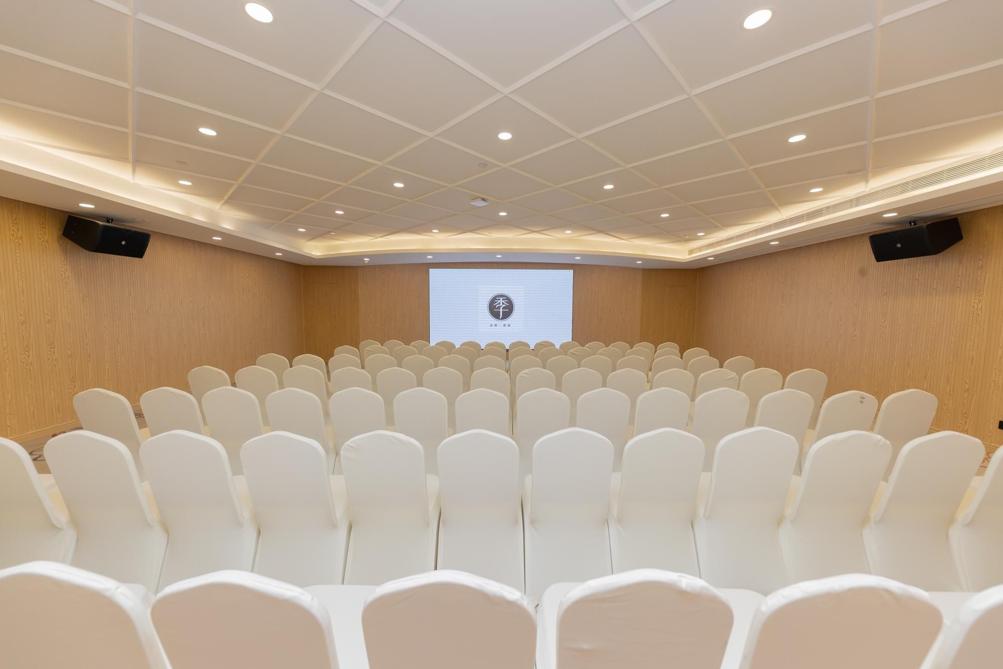Un salón de conferencias con filas de sillas blancas, una pared de madera, un techo con iluminación empotrada y una pantalla de proyección al frente.