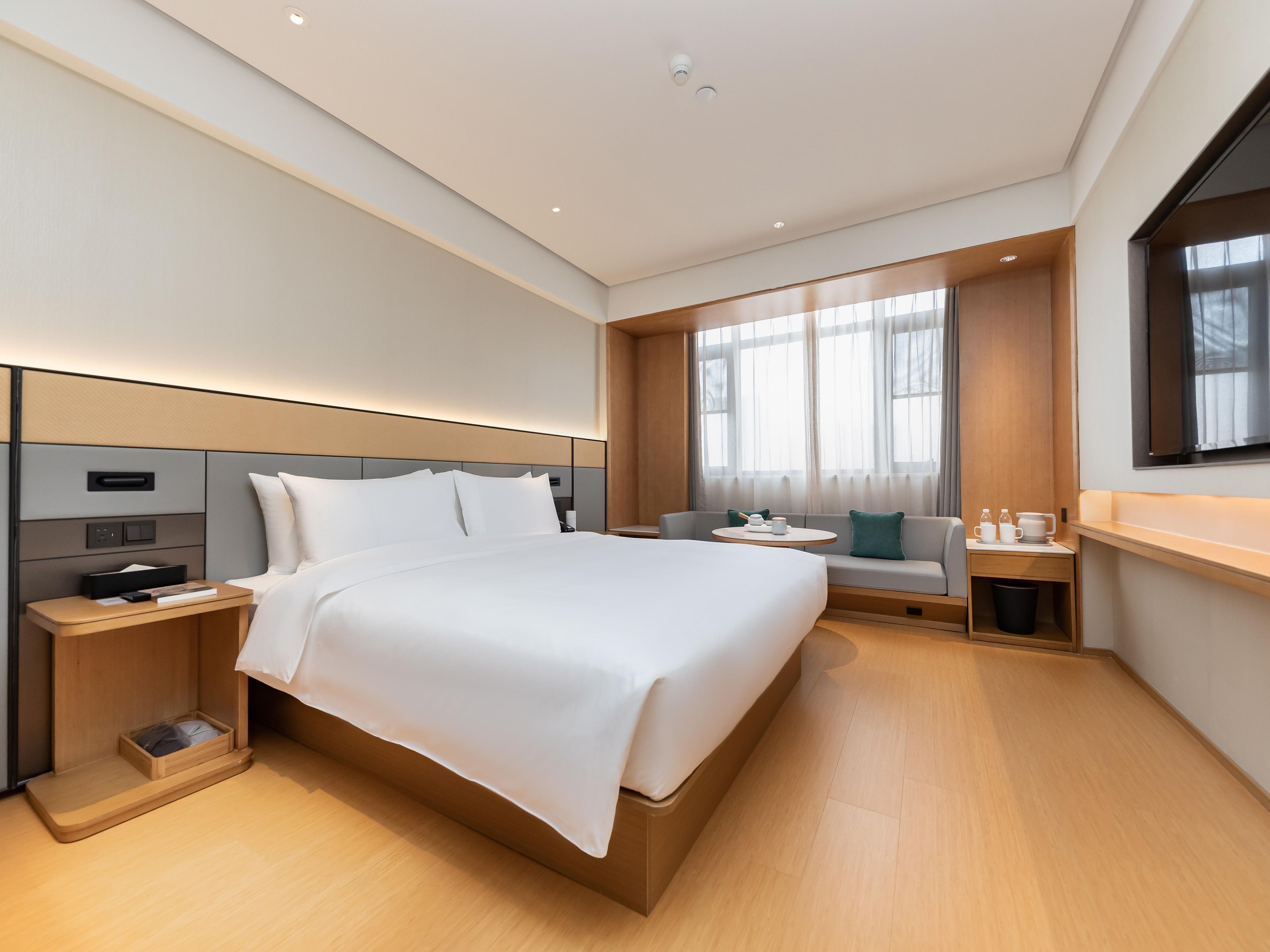 Una habitación de hotel moderna con una cama grande, mesitas de noche, un área de estar y un ventanal amplio.