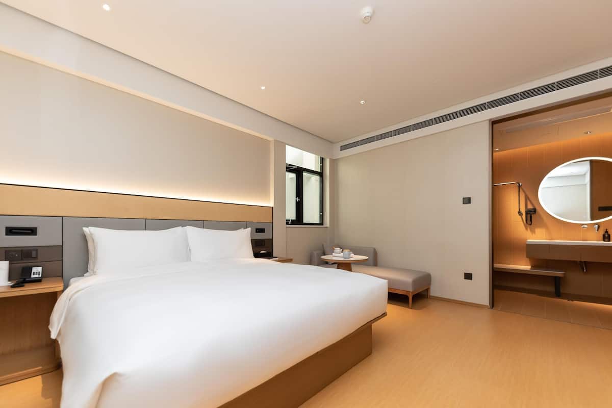 Una moderna camera d'albergo con un letto grande, comodini, uno specchio rotondo e un piccolo angolo salotto.