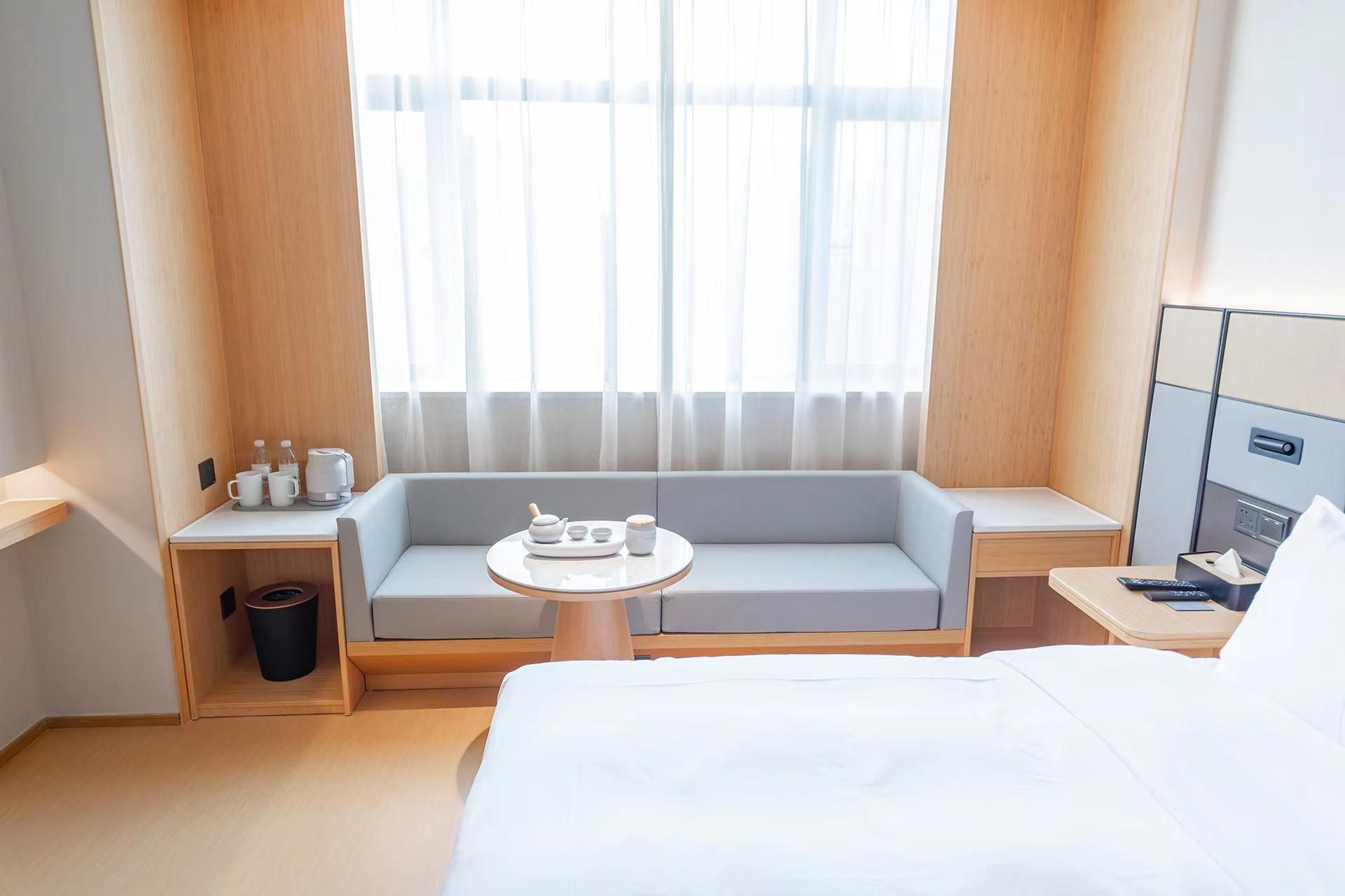 Una habitación de hotel con cama, una pequeña mesa redonda, un sofá, una ventana con cortinas traslúcidas y una mesita de noche con lámpara y control remoto.