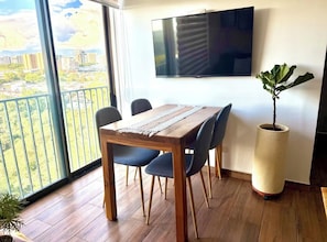 A dining area with a wooden table, four chairs, a large potted plant, a flat-screen TV on the wall, and a balcony with a city view.