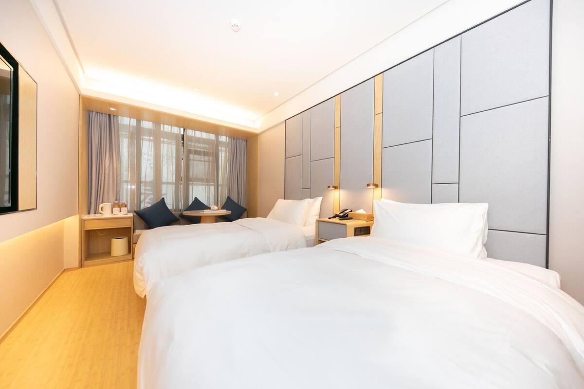 Habitación de hotel con dos camas, un ventanal con cortinas, una mesita de noche con lámpara y un panel decorativo en la pared.