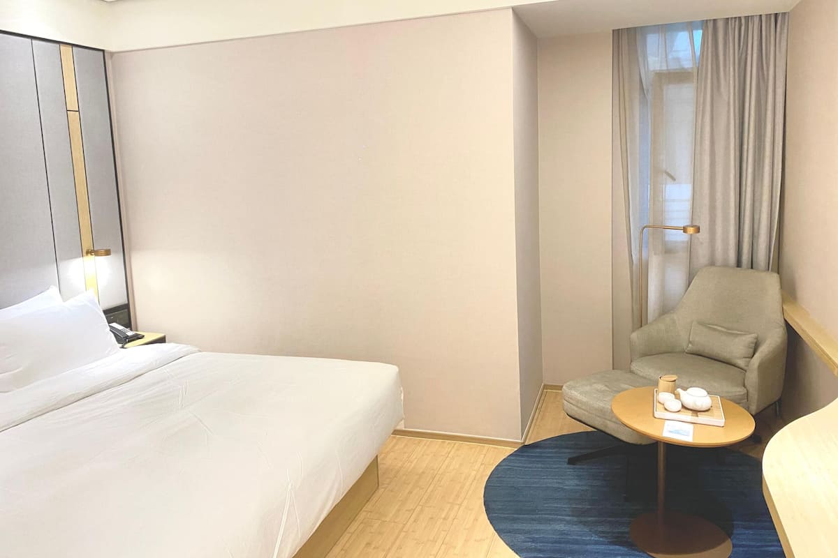 Una habitación de hotel con una cama, una silla, una mesita con tetera y una lámpara.