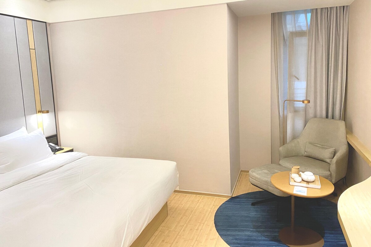 Una habitación de hotel con una cama, una silla, una mesita con tetera y una lámpara.