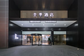Entrada de hotel com porta giratória, placa indicando 'JI HOTEL' e acesso visível ao saguão do hotel.