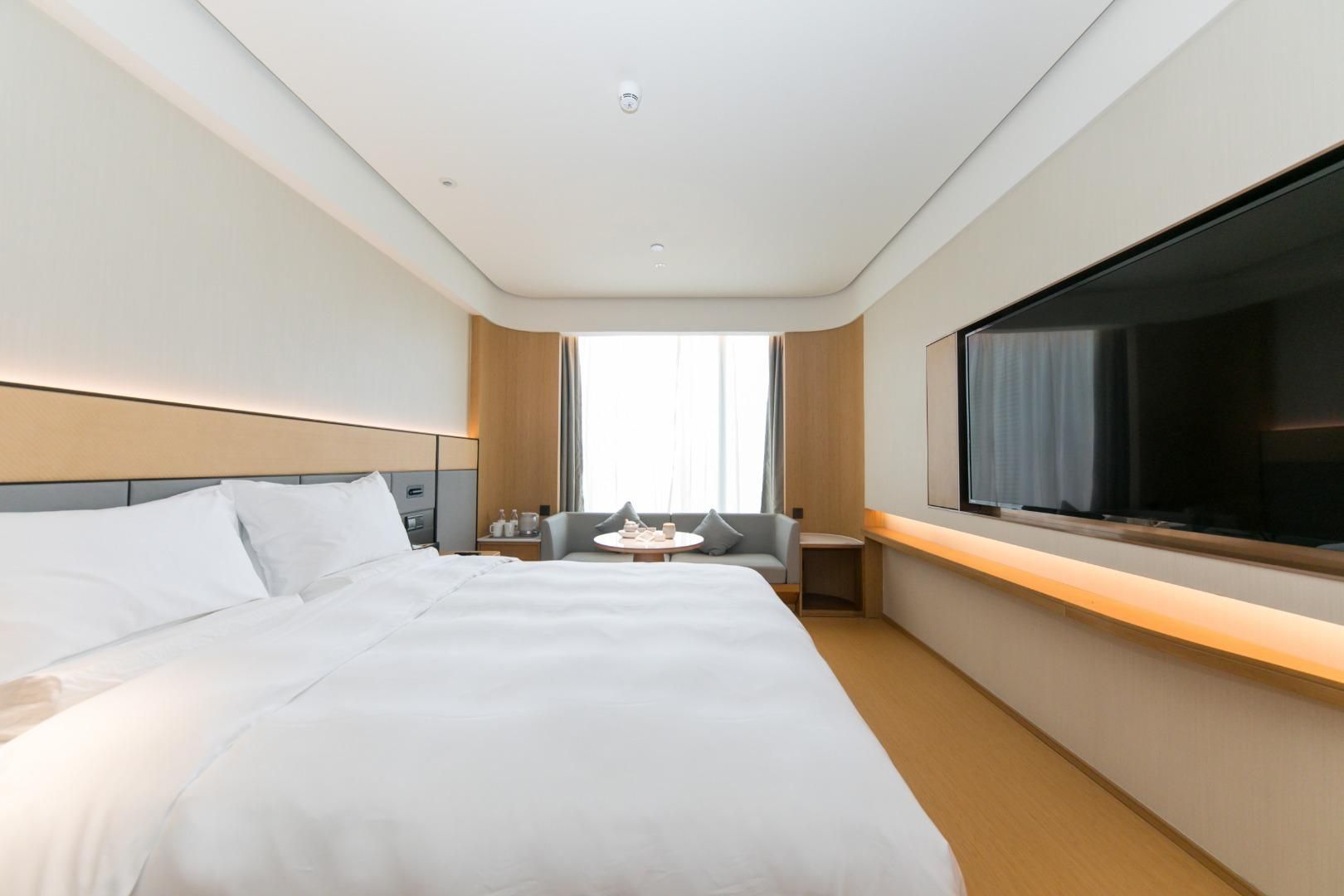 Quarto de hotel com uma cama grande, televisão fixada na parede, uma mesinha com uma cadeira e uma janela com cortinas.