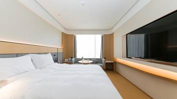 Quarto de hotel com uma cama grande, televisão fixada na parede, uma mesinha com uma cadeira e uma janela com cortinas.