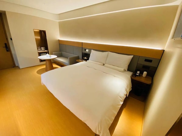 A hotel room with a large bed, a bedside table, a chair, a small round table, and a built-in shelf. - Ji Hotel (Nanjing)