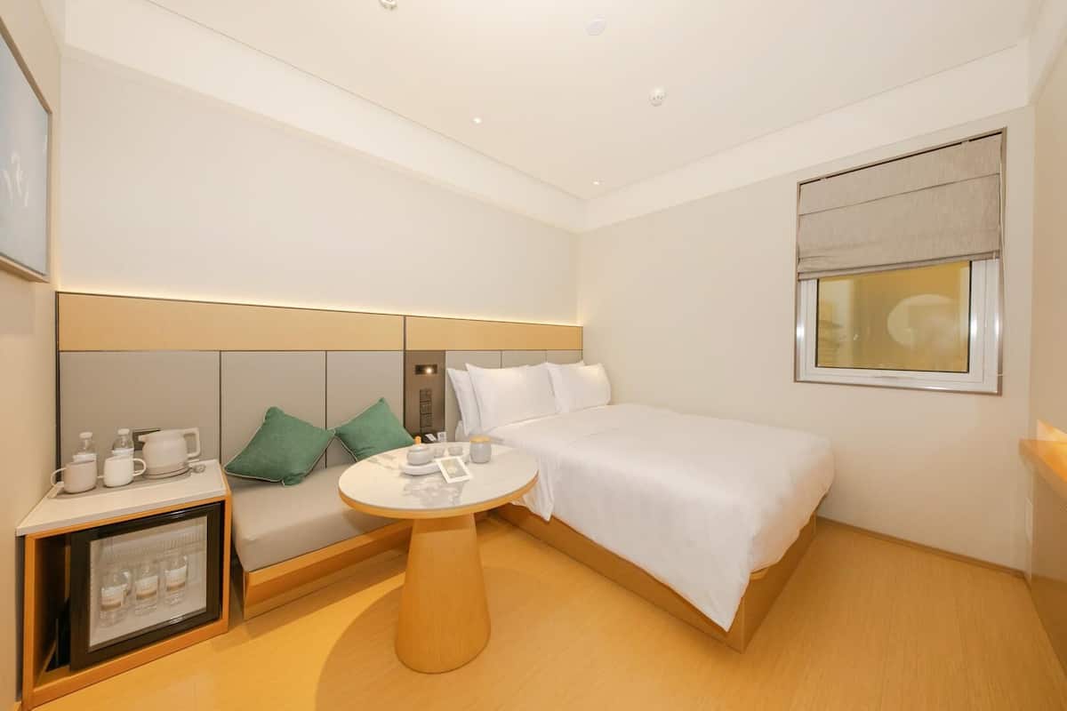 A hotel room with a bed, a bedside table with a lamp, a small seating area with cushions, a minibar, and a window with blinds.