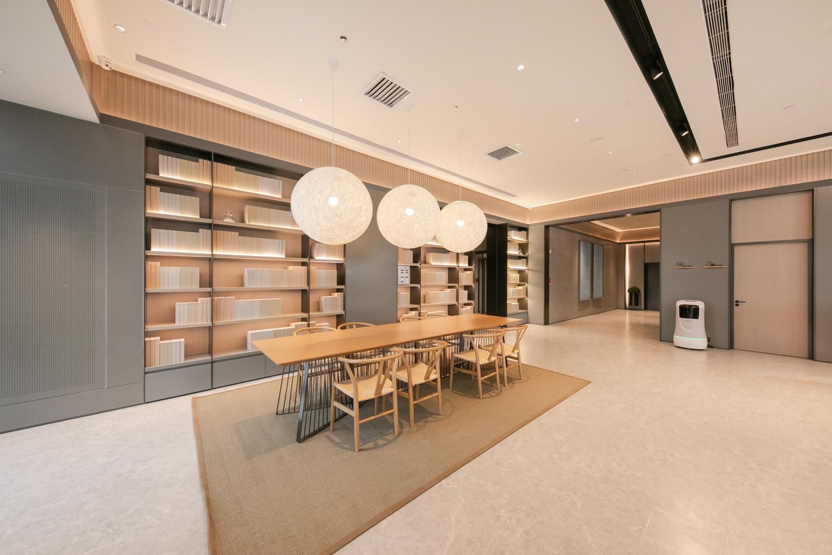 Una biblioteca amplia y moderna con una larga mesa de madera y sillas, rodeada de estanterías y lámparas colgantes.