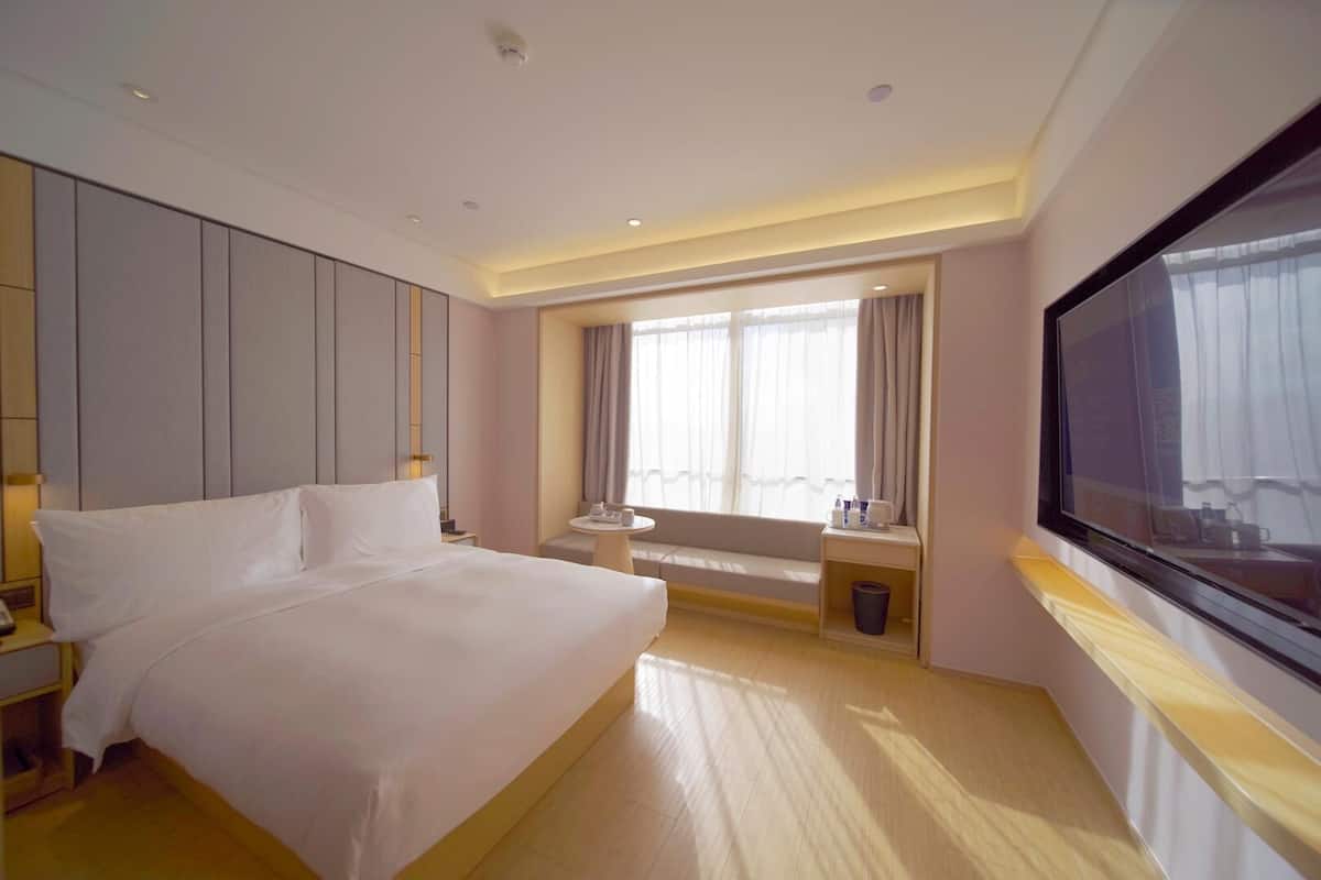 Una habitación de hotel con una cama grande, televisión, ventana con cortinas, una mesita pequeña y un banco.
